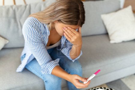 Photo for Sad, worried woman checking her recent pregnancy test, sitting on couch at home. Maternity, child birth and family problems concept. unwanted pregnancy - Royalty Free Image