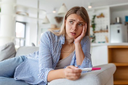 Photo for Sad, worried woman checking her recent pregnancy test, sitting on couch at home. Maternity, child birth and family problems concept. unwanted pregnancy - Royalty Free Image