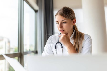 Smiling professional female doctor wearing uniform taking notes in medical journal, filling documents, patient illness history, looking at laptop screen, student watching webinar