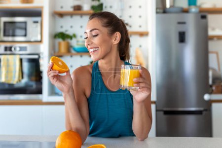 Foto de Hermosa joven bebiendo jugo de naranja fresco en la cocina. Dieta saludable. Feliz joven con vaso de jugo y naranja en la mesa en la cocina. - Imagen libre de derechos