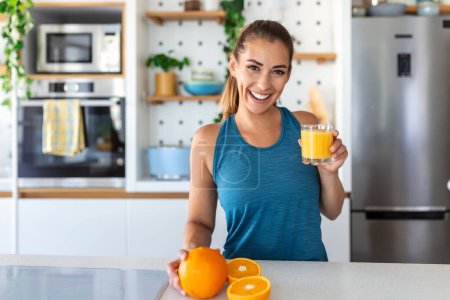 Photo for Beautiful young woman drinking fresh orange juice in kitchen. Healthy diet. Happy young woman with glass of juice and orange at table in kitchen. - Royalty Free Image