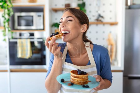 Photo for Young woman making pancakes at kitchen. Young housewife enjoying blueberry pancakes for breakfast - Royalty Free Image