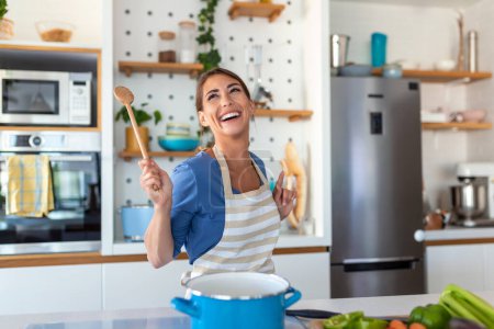Excited woman singing and dancing in modern kitchen at home, happy woman holding spatula as microphone, dancing, listening to music, having fun with kitchenware, preparing breakfast