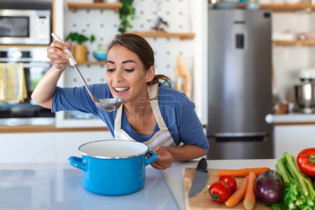 Photo for Happy Young Woman Cooking Tasting Dinner In A Pot Standing In Modern Kitchen At Home. Housewife Preparing Healthy Food Smiling . Household And Nutrition. Dieting Recipes Concept - Royalty Free Image