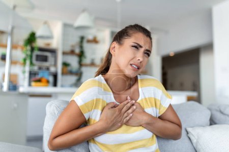 Téléchargez les photos : Stressé jeune femme se sentant douleur et toucher la poitrine souffrent de maladie du cœur à la maison tout en ayant une crise cardiaque, infarctus - en image libre de droit