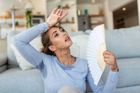 Photo for Woman puts head on sofa cushions feels sluggish due unbearable heat, waves hand fan cool herself, hot summer flat without air-conditioner climate control system concept - Royalty Free Image