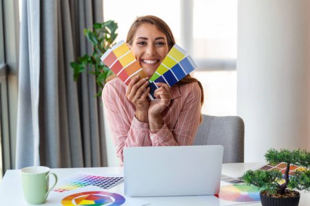 Téléchargez les photos : Joyeux jeune femme designer ayant une vidéoconférence avec les clients, assis au bureau devant l'ordinateur, tenant des palettes de couleurs, geste et sourire, espace de copie - en image libre de droit