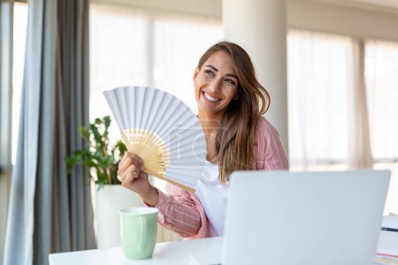 Photo for Tired overheated young woman hold wave fan suffer from heat sweating indoor work on laptop at home office, annoyed woman feel uncomfortable hot summer weather problem no air conditioner concept - Royalty Free Image