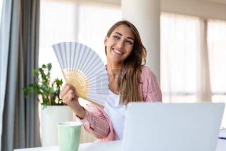 Photo for Tired overheated young woman hold wave fan suffer from heat sweating indoor work on laptop at home office, annoyed woman feel uncomfortable hot summer weather problem no air conditioner concept - Royalty Free Image