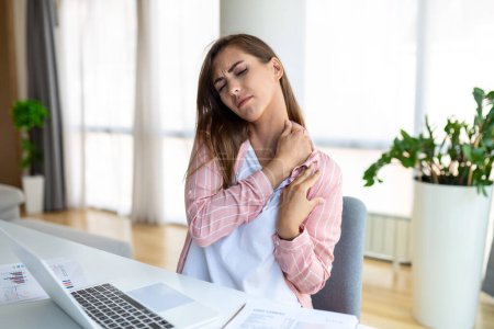 Foto de Retrato de una joven mujer estresada sentada en el escritorio de la oficina en casa delante de la computadora portátil, tocando el hombro dolorido con expresión dolorida, sufriendo de dolor de hombro después de trabajar en la computadora portátil - Imagen libre de derechos