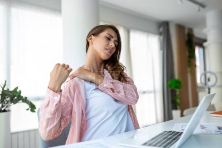 Téléchargez les photos : Portrait d'une jeune femme stressée assise au bureau à la maison devant un ordinateur portable, touchant l'épaule douloureuse avec une expression douloureuse, souffrant de douleurs à l'épaule après avoir travaillé sur un ordinateur portable - en image libre de droit
