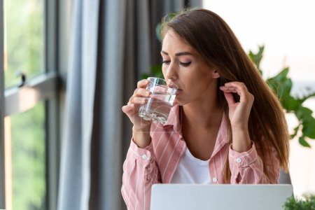 Foto de Dolor de cabeza. Foto de cerca de una mujer joven, que está sentada en la oficina, hidratándose, bebiendo agua. - Imagen libre de derechos