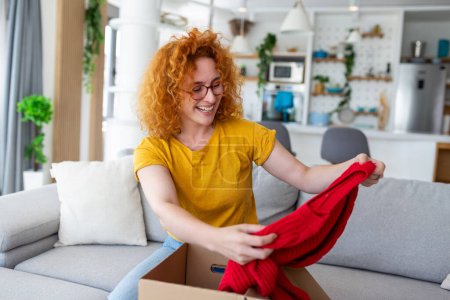 Foto de Hermosa joven alegre pelo rojo mujer unboxing un paquete en la sala de estar de un nuevo suéter. - Imagen libre de derechos