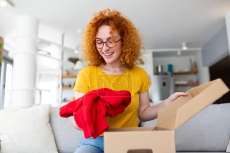 Foto de Hermosa joven alegre pelo rojo mujer unboxing un paquete en la sala de estar de un nuevo suéter. - Imagen libre de derechos