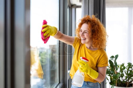 Photo for Housework and housekeeping concept - happy woman in gloves cleaning window with rag and cleanser spray at home - Royalty Free Image