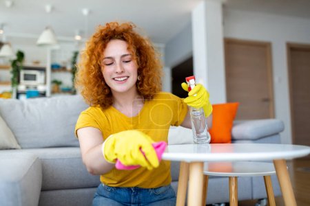 Téléchargez les photos : Beautiful young woman cleaning the house. Girl rubs dust. Smiling woman wearing rubber protective yellow gloves cleaning with rag and spray bottle detergent. Home, housekeeping concept. - en image libre de droit