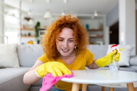 Téléchargez les photos : Beautiful young woman cleaning the house. Girl rubs dust. Smiling woman wearing rubber protective yellow gloves cleaning with rag and spray bottle detergent. Home, housekeeping concept. - en image libre de droit