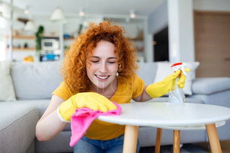 Foto de Beautiful young woman cleaning the house. Girl rubs dust. Smiling woman wearing rubber protective yellow gloves cleaning with rag and spray bottle detergent. Home, housekeeping concept. - Imagen libre de derechos