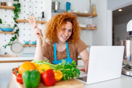 Téléchargez les photos : Une jeune femme apprend à cuisiner, elle regarde des recettes vidéo sur un ordinateur portable dans la cuisine et cuisine un plat. Concept de cuisine à la maison - en image libre de droit