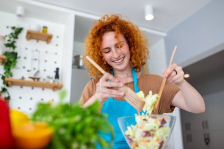 Foto de Hermosa linda joven sonriente mujer en la cocina está preparando una ensalada vegana en ropa casual
. - Imagen libre de derechos
