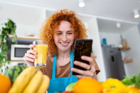 Foto de Mujer bonita sonriente mirando el teléfono móvil y sosteniendo el vaso de jugo de naranja mientras cocina verduras frescas en el interior de la cocina en casa - Imagen libre de derechos