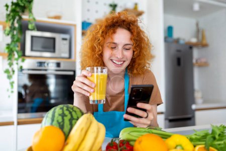 Photo for Smiling pretty woman looking at mobile phone and holding glass of orange juice while cooking fresh vegetables in kitchen interior at home - Royalty Free Image