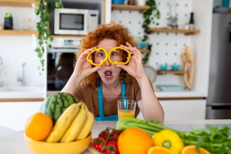 Foto de Foto de mujer joven sonriendo y sosteniendo círculos de pimienta en sus ojos mientras cocina ensalada con verduras frescas en el interior de la cocina en casa - Imagen libre de derechos
