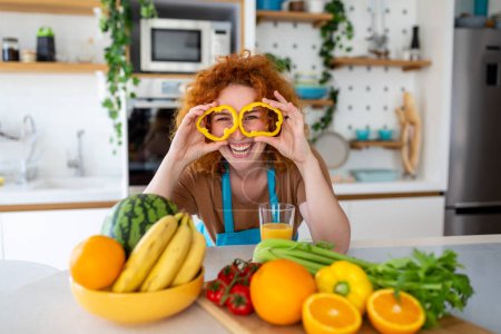Photo for Photo of young woman smiling and holding pepper circles on her eyes while cooking salad with fresh vegetables in kitchen interior at home - Royalty Free Image