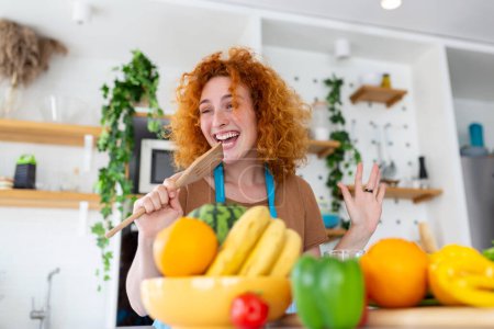 Photo for Funny beautiful woman singing into spatula, cooking in modern kitchen, holding spatula as microphone, dancing, listening to music, playful girl having fun with kitchenware, preparing food. - Royalty Free Image