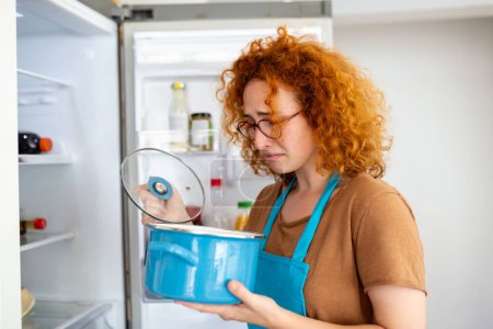 Foto de Bad Food In Fridge, young woman in holding her nose because of bad smell from food in refrigerator at home - Imagen libre de derechos