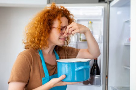 Photo for Bad Food In Fridge, young woman in holding her nose because of bad smell from food in refrigerator at home - Royalty Free Image