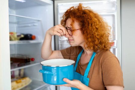 Photo for Bad Food In Fridge, young woman in holding her nose because of bad smell from food in refrigerator at home - Royalty Free Image