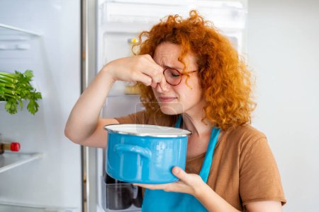 Téléchargez les photos : Bad Food In Fridge, young woman in holding her nose because of bad smell from food in refrigerator at home - en image libre de droit
