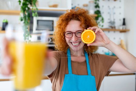 Téléchargez les photos : Photo de mignonne femme caucasienne souriant et tenant la partie orange et le jus d'orange dans l'intérieur de la cuisine à la maison - en image libre de droit
