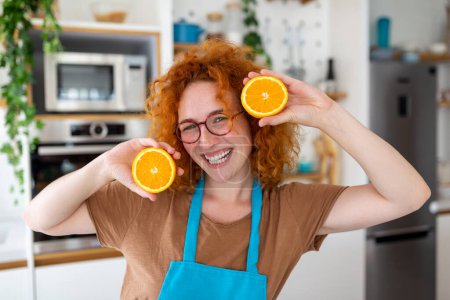 Foto de Foto de linda mujer caucásica sonriendo y sosteniendo dos partes de naranja mientras cocina ensalada de verduras en el interior de la cocina en casa - Imagen libre de derechos