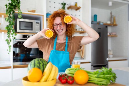 Foto de Foto de linda mujer caucásica sonriendo y sosteniendo dos partes de naranja mientras cocina ensalada de verduras en el interior de la cocina en casa - Imagen libre de derechos