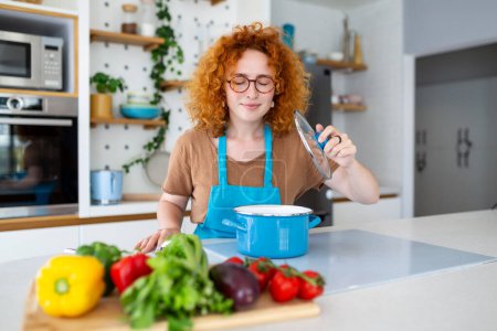 Cheerful young pretty female in apron prepare lunch and smell dish in kitchen at home. Woman cooking dinner for family at new recipe at home.