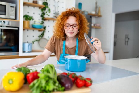 Photo for Cheerful young pretty female in apron prepare lunch and smell dish in kitchen at home. Woman cooking dinner for family at new recipe at home. - Royalty Free Image