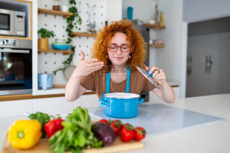 Photo for Cheerful young pretty female in apron prepare lunch and smell dish in kitchen at home. Woman cooking dinner for family at new recipe at home. - Royalty Free Image