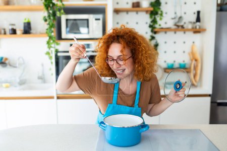 Photo for Young smiling woman enjoying in taste and smell while cooking lunch in the kitchen. - Royalty Free Image