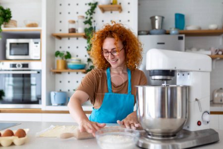 Photo for Happy attractive young adult woman housewife baker wear apron holding pin rolling dough on kitchen table baking pastry concept cooking cake biscuit doing bakery making homemade pizza at home - Royalty Free Image