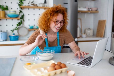 Téléchargez les photos : Professionnel belle jeune femme heureuse blogue pour son canal de cuisine sur la vie saine dans la cuisine de sa maison et regardant la caméra sur un ordinateur portable - en image libre de droit