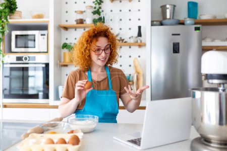 Téléchargez les photos : Professionnel belle jeune femme heureuse blogue pour son canal de cuisine sur la vie saine dans la cuisine de sa maison et regardant la caméra sur un ordinateur portable - en image libre de droit