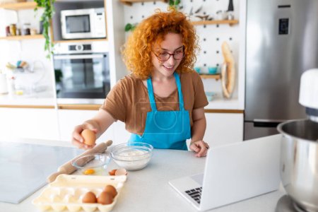 Photo for Professional beautiful happy young woman is blogging for her kitchen channel about healthy living in the kitchen of her home and looking on camera on a laptop - Royalty Free Image