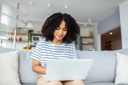 Téléchargez les photos : Jolie jeune femme assise seule sur son canapé à la maison et utilisant son ordinateur portable - en image libre de droit