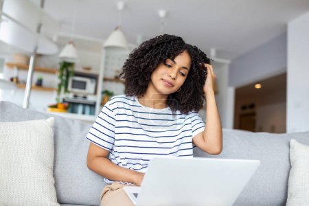 Photo for Smiling young woman freelancer working from home, sitting on couch, using laptop, copy space. Relaxed african american lady enjoying her weekend, surfing on internet on laptop - Royalty Free Image