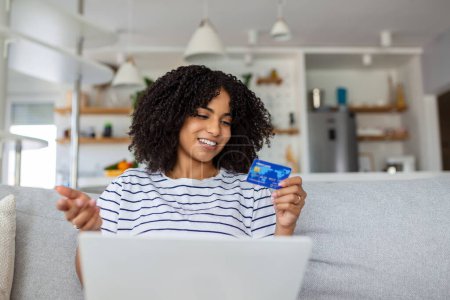 Foto de Mujer joven en el sofá de compras en línea con tarjeta de débito. Hermosa mujer africana usando ordenador portátil para compras en línea en casa - Imagen libre de derechos