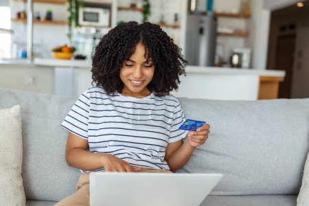 Foto de Mujer joven en el sofá de compras en línea con tarjeta de débito. Hermosa mujer africana usando ordenador portátil para compras en línea en casa - Imagen libre de derechos