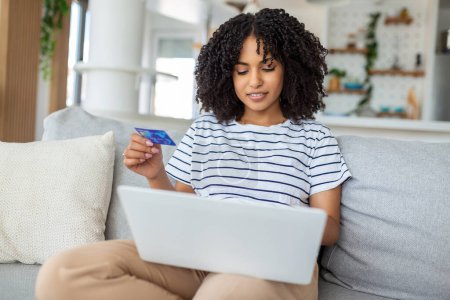 Photo for Young Woman On Sofa Shopping Online With Debit Card. Beautiful girl using laptop computer for online shopping at home - Royalty Free Image