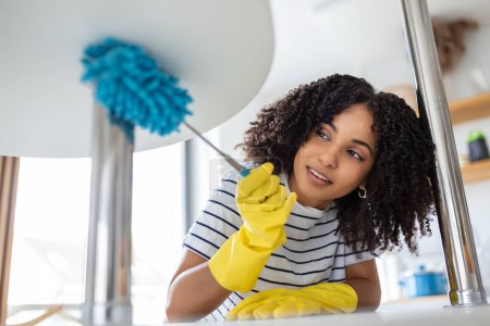 Photo for Young housekeeper in apron dusting the shelf by duster carefully cleaning in living room at home. young wife in rubber gloves doing housework. - Royalty Free Image